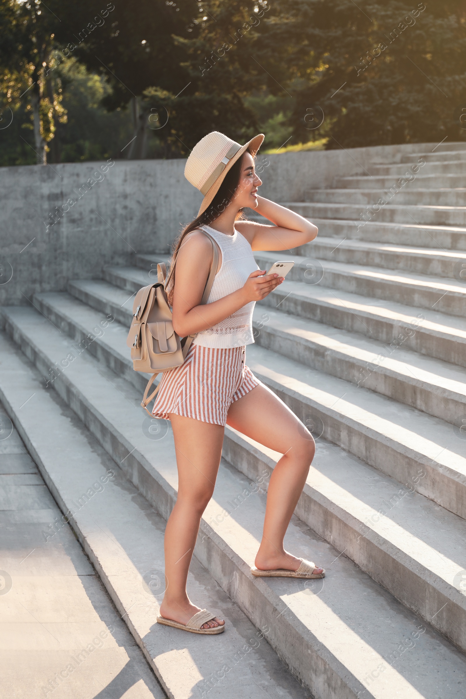 Photo of Beautiful young woman with stylish backpack and smartphone walking up stairs outdoors