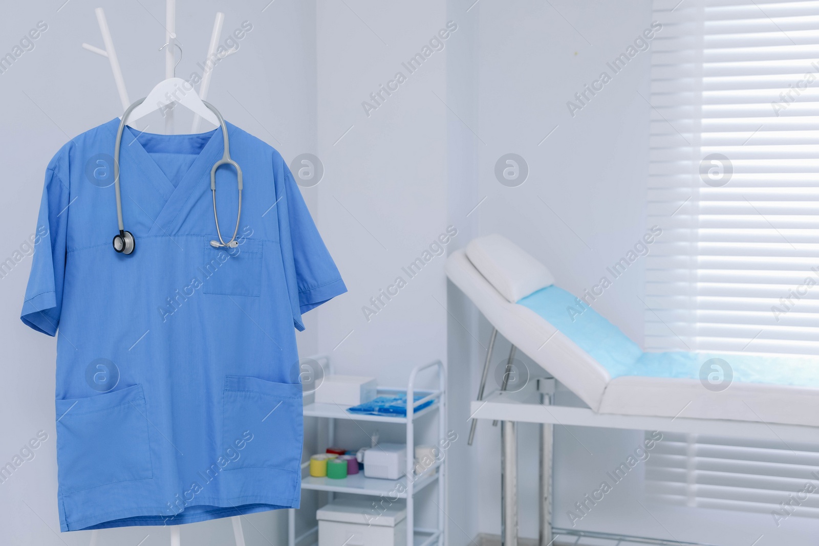 Photo of Blue medical uniform and stethoscope hanging on rack in clinic. Space for text