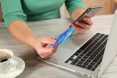 Online payment. Woman using credit card and smartphone near laptop at light wooden table indoors, closeup