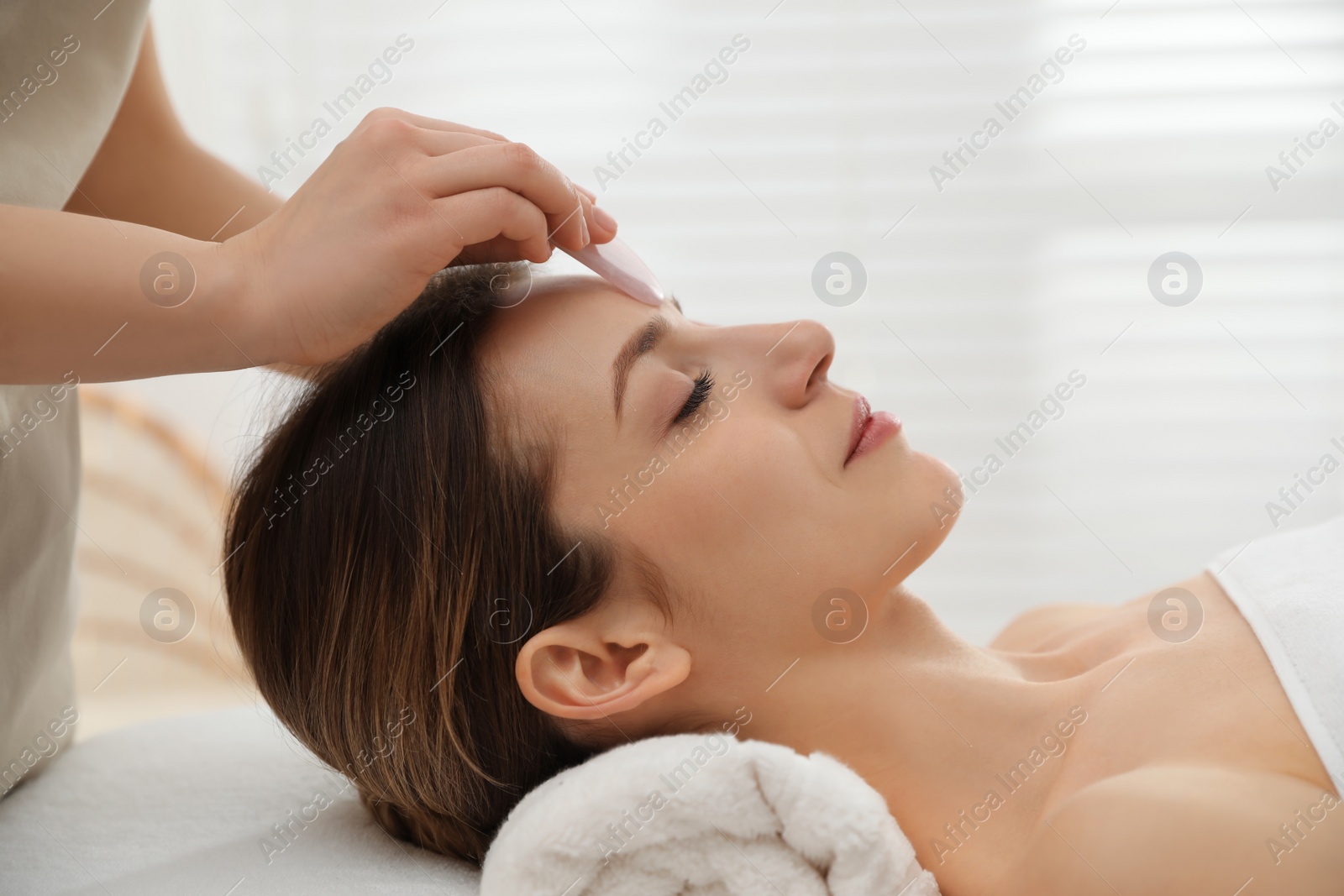 Photo of Young woman receiving facial massage with gua sha tool in beauty salon