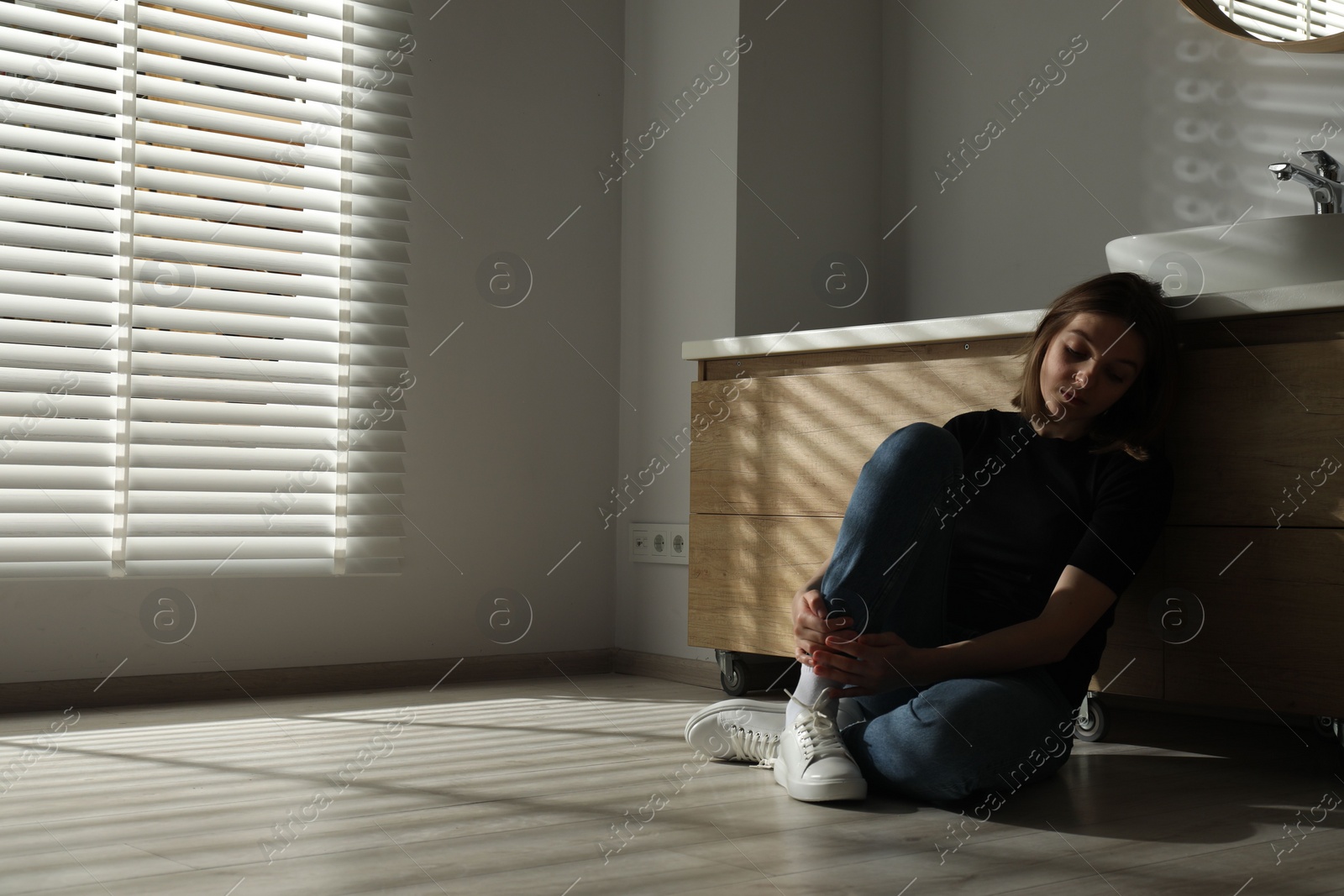 Photo of Sad young woman sitting on floor in bathroom, space for text