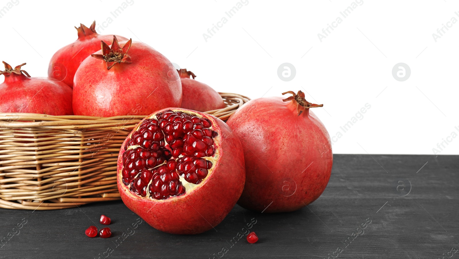 Photo of Fresh pomegranates in wicker basket on black wooden table against white background, space for text