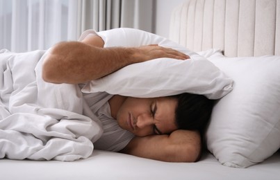 Handsome man sleeping under pillow in bed at home