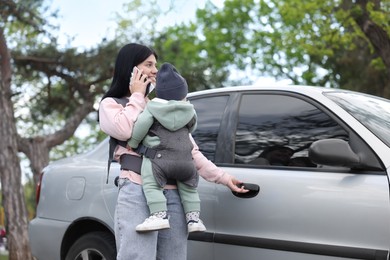 Photo of Mother holding her child in sling (baby carrier) while talking on smartphone near car outdoors