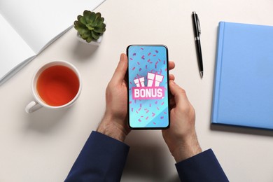 Image of Bonus gaining. Man holding smartphone at white table, top view. Illustration of gift boxes, word and falling confetti on device screen