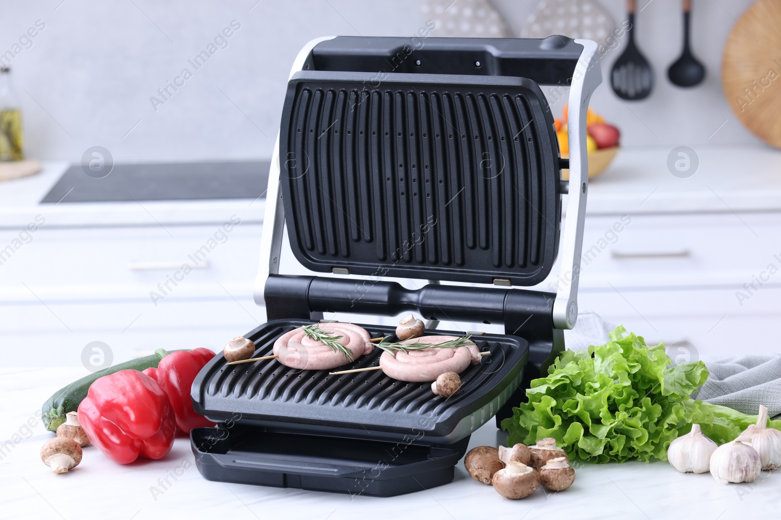 Photo of Electric grill with homemade sausages, rosemary and vegetables on white table in kitchen