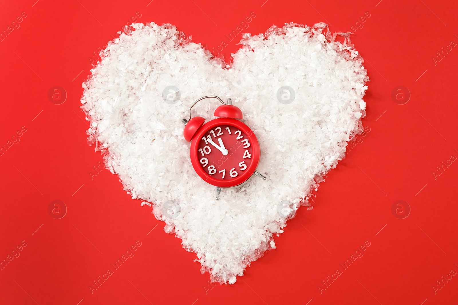 Photo of Alarm clock in heart shaped pile of snow on red background, flat lay. New Year countdown