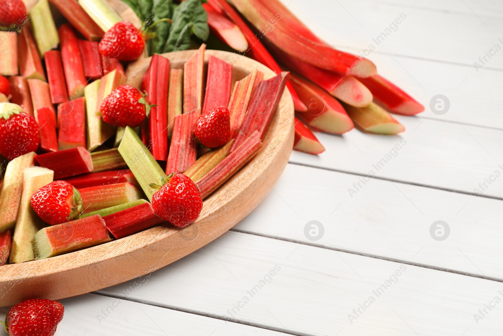 Photo of Cut fresh rhubarb stalks and strawberries on white wooden table, space for text