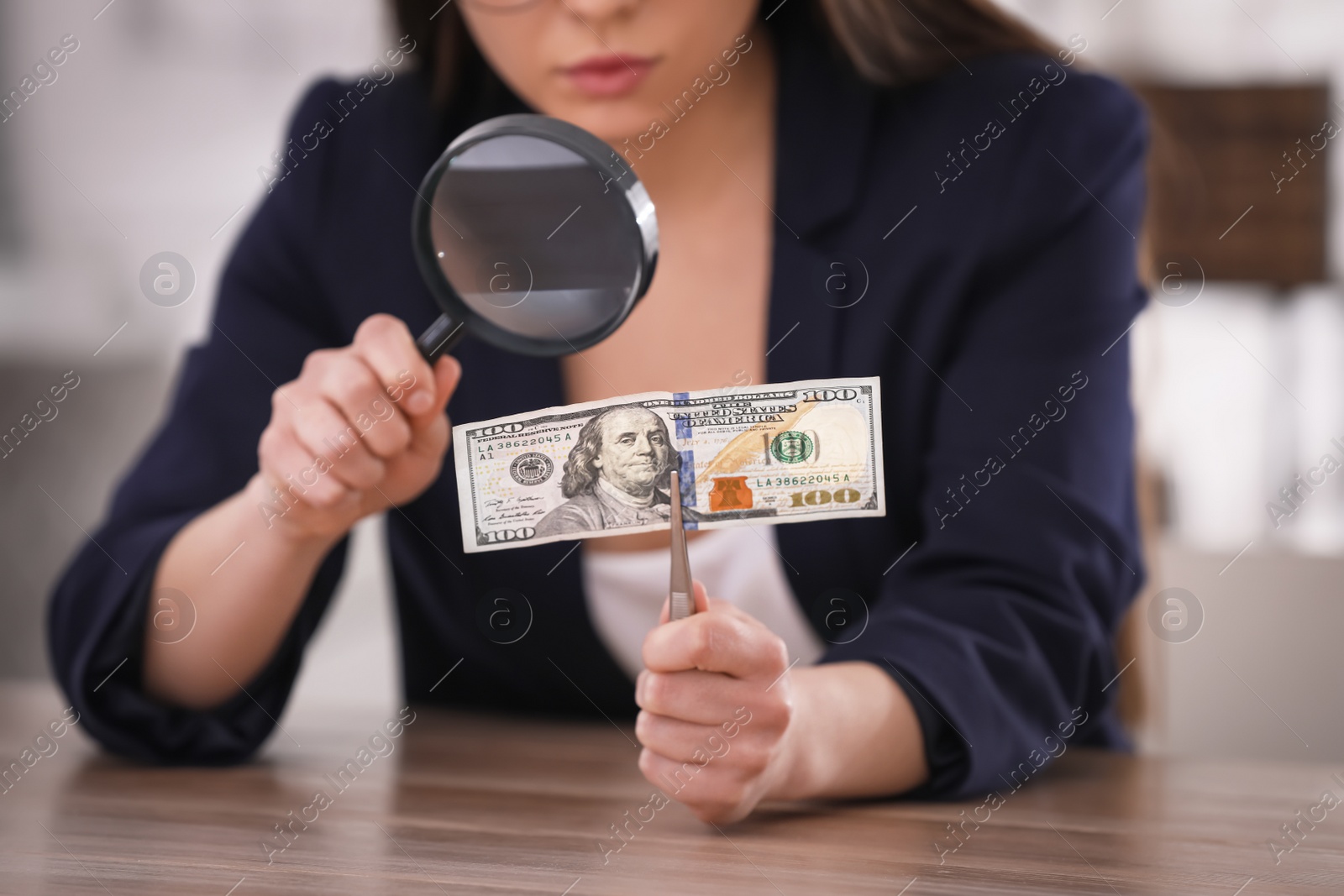 Photo of Expert authenticating 100 dollar banknote with magnifying glass at table in office, closeup. Fake money concept