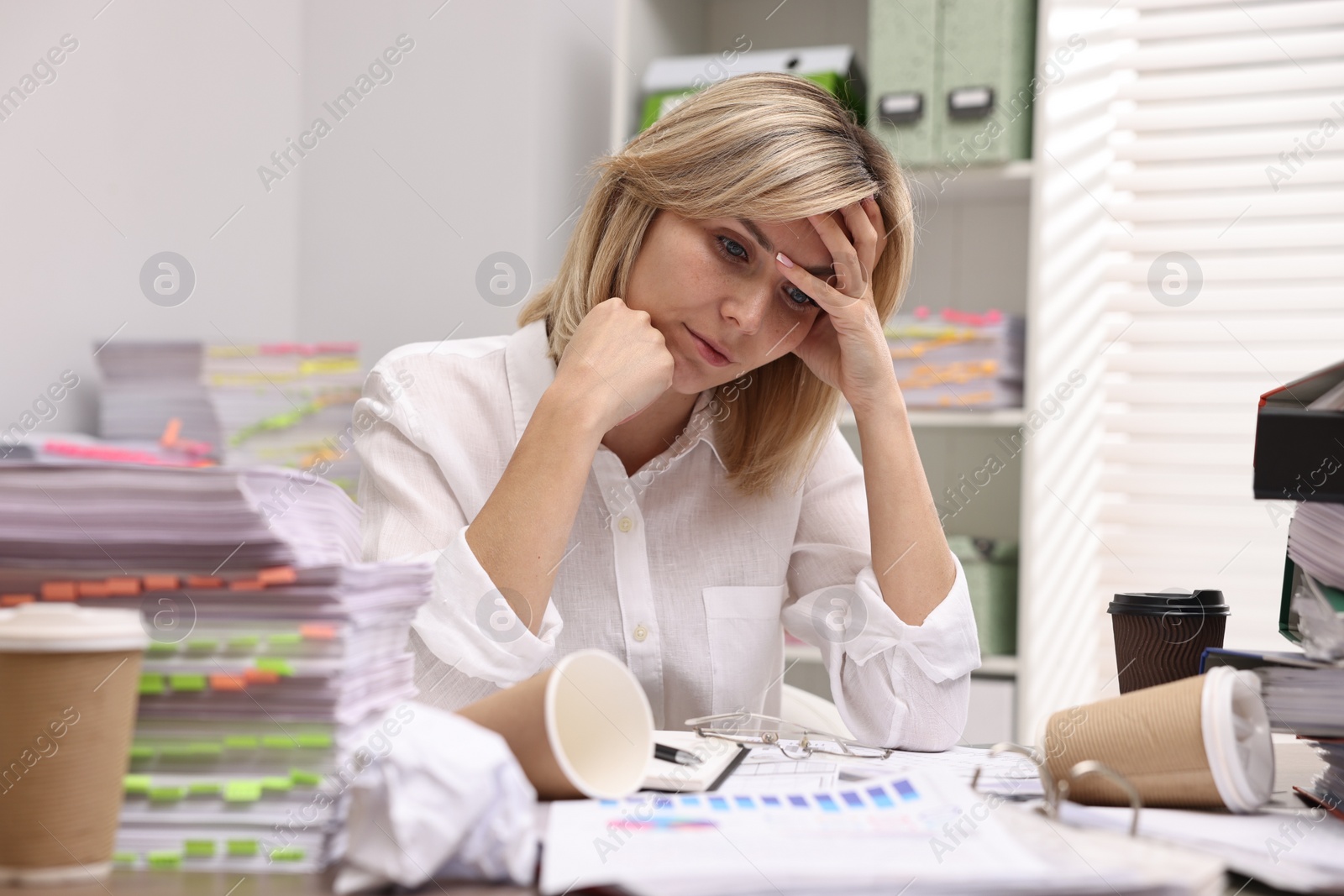 Photo of Overwhelmed woman surrounded by documents and paper coffee cups at workplace in office