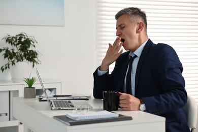 Photo of Sleepy man with cup of drink yawning at table in office