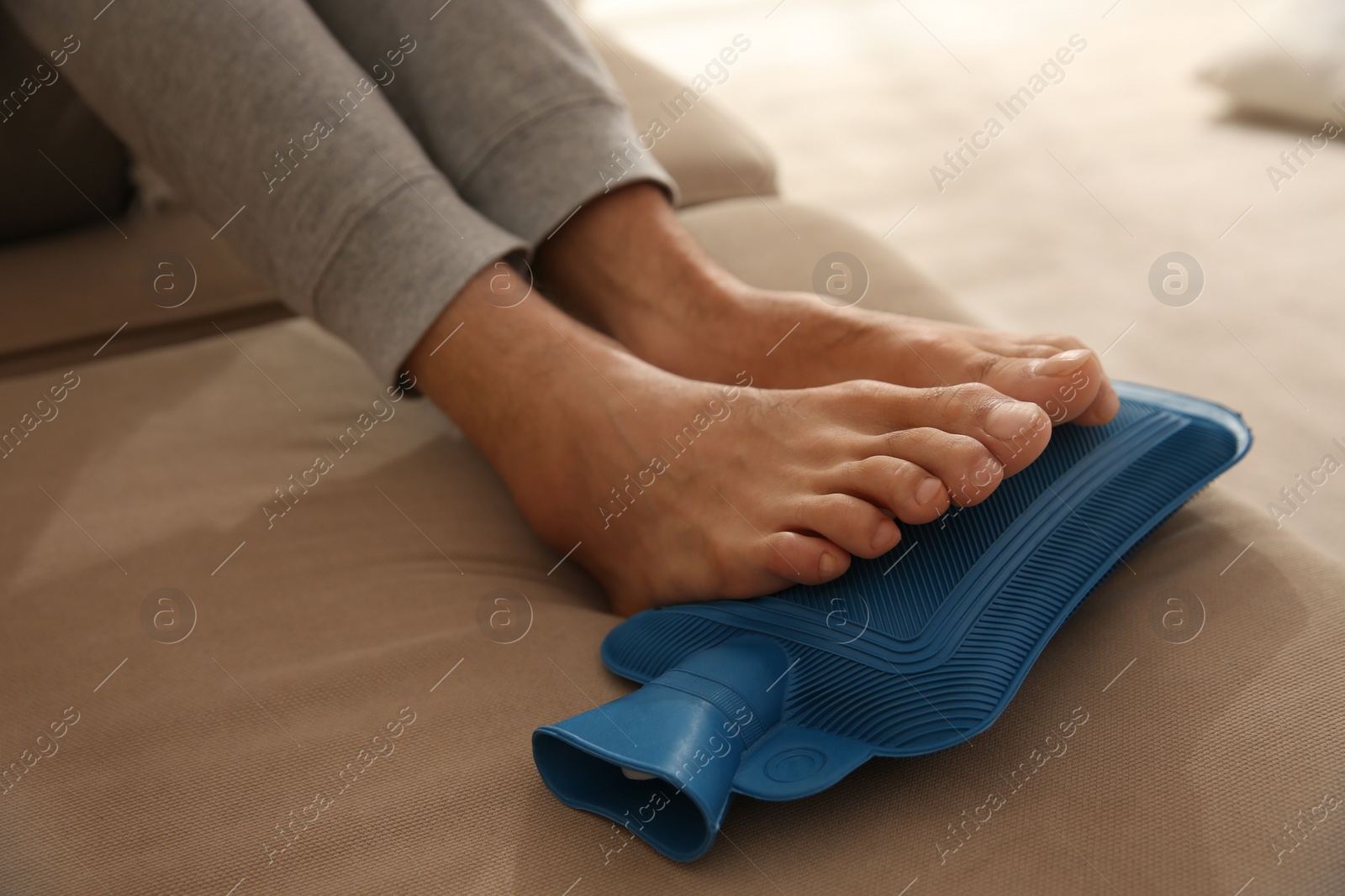 Photo of Man warming feet with hot water bottle on sofa, closeup