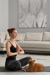 Beautiful woman with cute red cat practicing yoga on mat at home