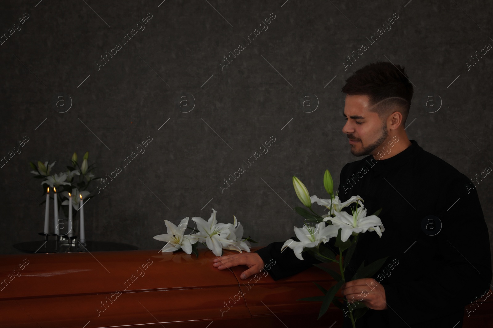 Photo of Sad young man with white lilies near casket in funeral home