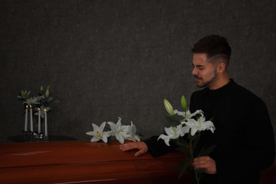 Photo of Sad young man with white lilies near casket in funeral home