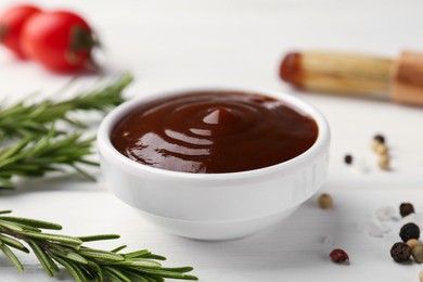Photo of Tasty barbeque sauce in bowl, rosemary and spices on white wooden table, closeup