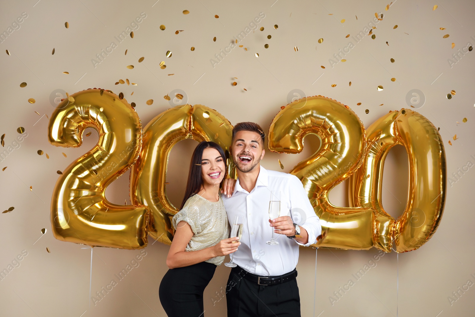 Photo of Happy young couple with glasses of champagne near golden 2020 balloons on beige background. New Year celebration