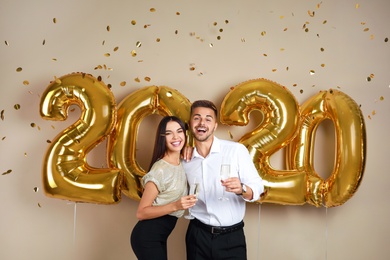 Happy young couple with glasses of champagne near golden 2020 balloons on beige background. New Year celebration