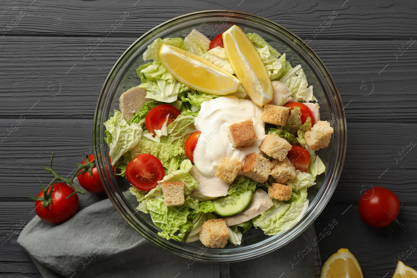 Photo of Bowl of delicious salad with Chinese cabbage, lemon, tomatoes and bread croutons on black wooden table, flat lay