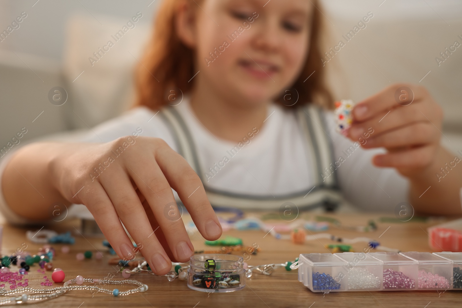 Photo of Cute girl making beaded jewelry at table in room, focus on hand