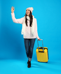 Young woman in warm clothes with suitcase on blue background. Winter vacation