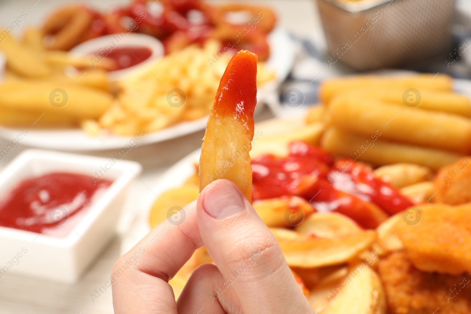 Photo of Woman holding delicious baked potato wedge with ketchup near different snacks at table, closeup