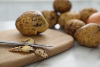 Photo of Fresh potatoes, peels, wooden board and knife on white marble table, closeup