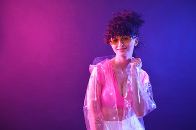 Photo of Beautiful young woman in transparent coat and sunglasses posing on color background in neon lights. Space for text