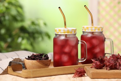 Photo of Refreshing hibiscus tea with ice cubes in mason jars and roselle flowers on wooden table against blurred green background