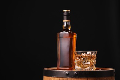 Photo of Whiskey in glasses and bottle on wooden barrel against black background, space for text