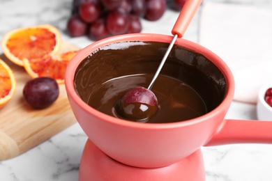 Photo of Dipping grape into pot with chocolate fondue on table, closeup