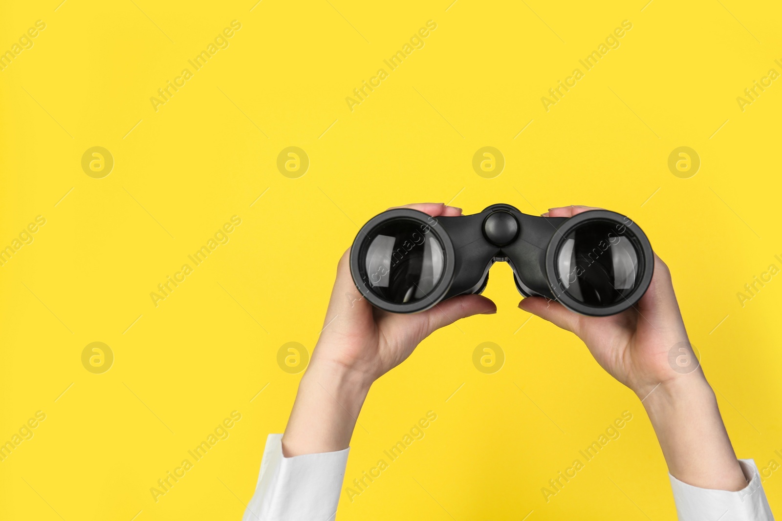 Photo of Woman holding modern binoculars on yellow background, closeup. Space for text