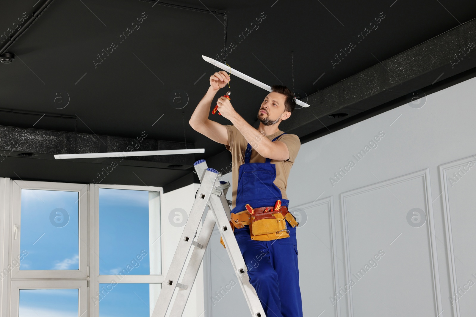 Photo of Electrician in uniform installing ceiling lamp indoors