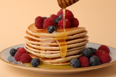 Photo of Pouring honey onto stack of tasty pancakes with raspberries and blueberries on pale orange background, closeup