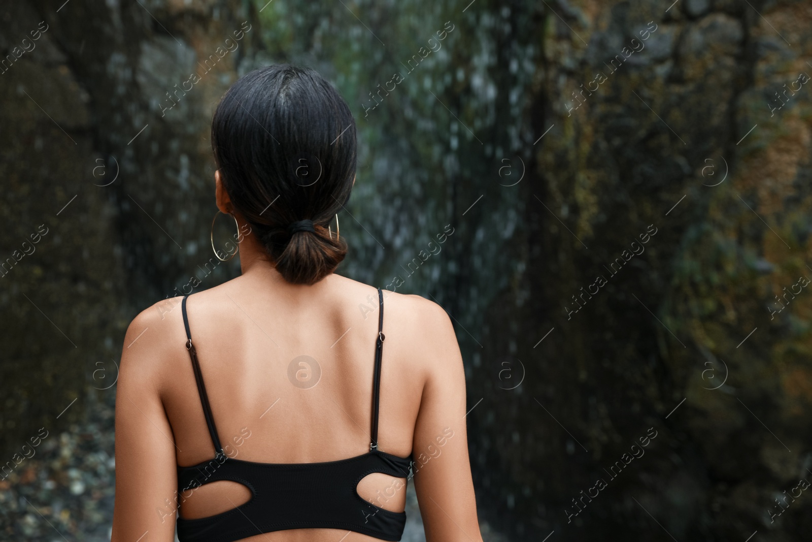 Photo of Young woman in stylish bikini near beautiful waterfall outdoors, back view. Space for text