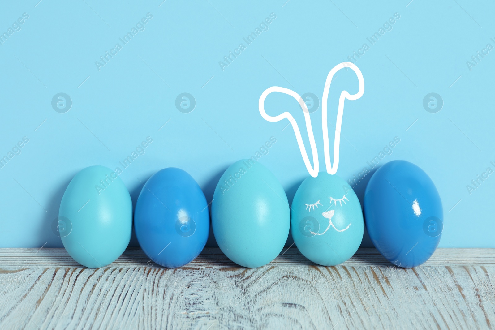 Image of One egg with drawn face and ears as Easter bunny among others on white wooden table against light blue background