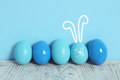 One egg with drawn face and ears as Easter bunny among others on white wooden table against light blue background