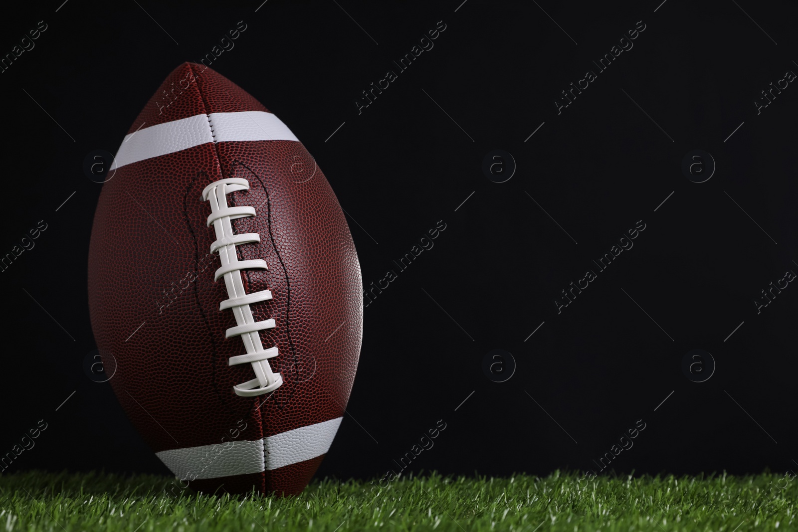 Photo of American football ball on green grass against black background. Space for text