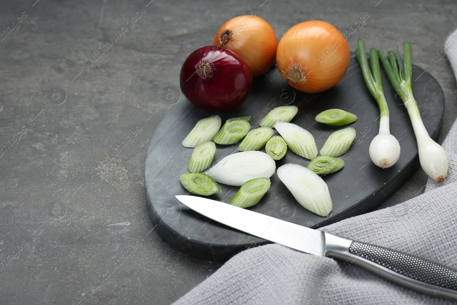 Photo of Board with different kinds of onions and knife on grey table, closeup. Space for text