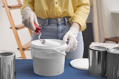 Photo of Woman dipping roller into bucket with paint at blue wooden table indoors, closeup