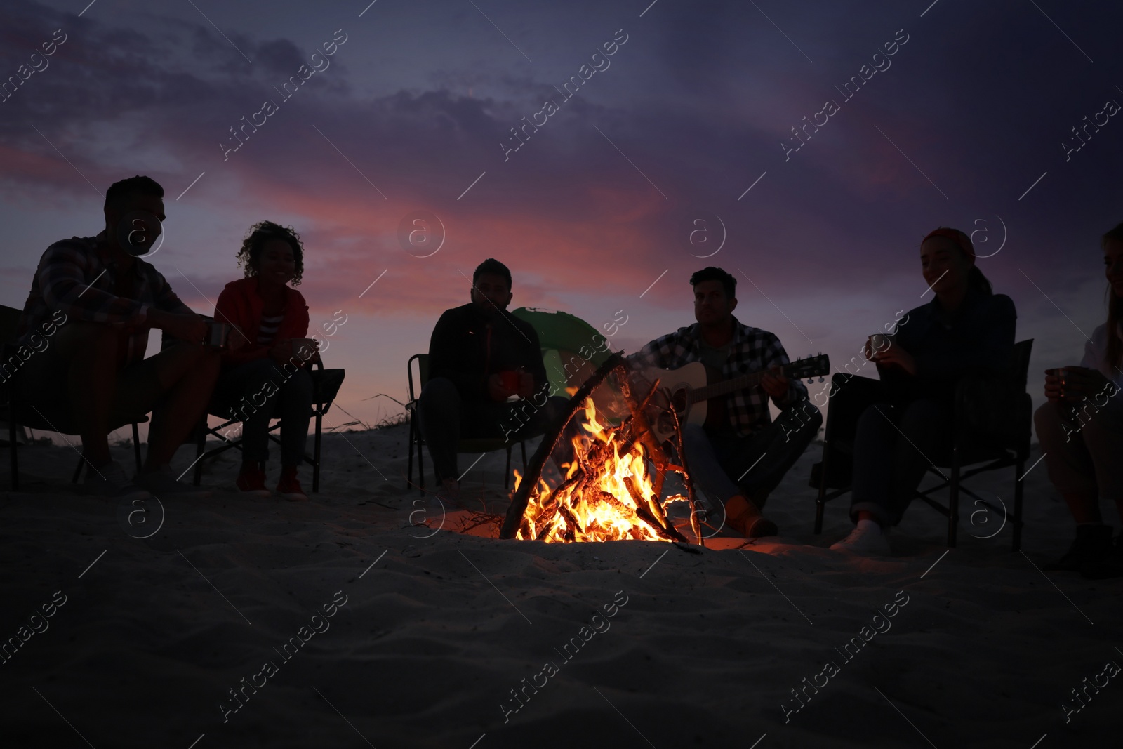 Photo of Group of friends gathering around bonfire in evening. Camping season