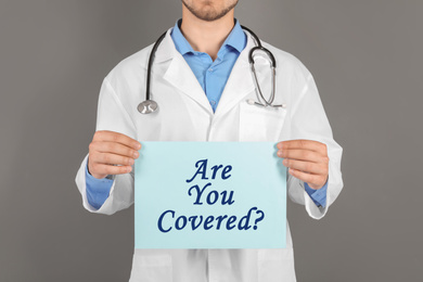 Image of Doctor holding paper with text ARE YOU COVERED? on grey background, closeup. Insurance concept