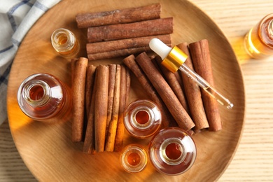 Photo of Bottles of essential oils and cinnamon sticks on wooden plate, top view