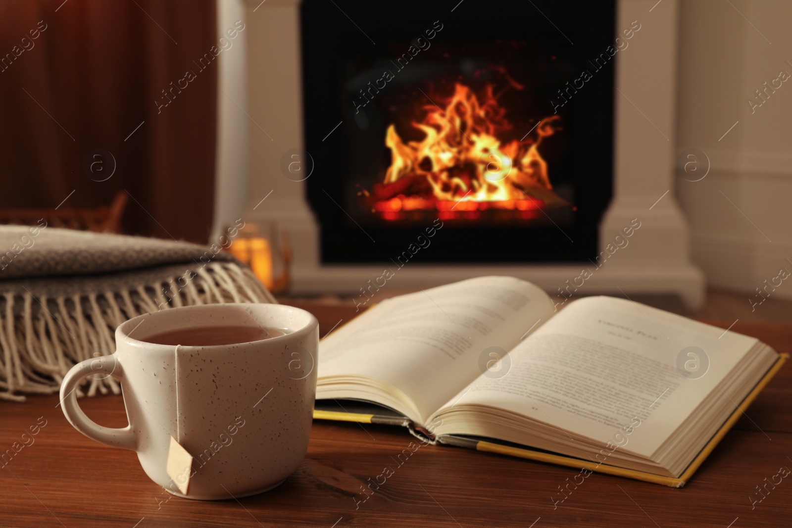 Photo of Cup of hot tea and book on wooden table near fireplace at home. Cozy atmosphere