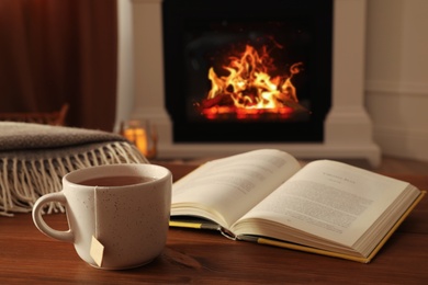 Photo of Cup of hot tea and book on wooden table near fireplace at home. Cozy atmosphere