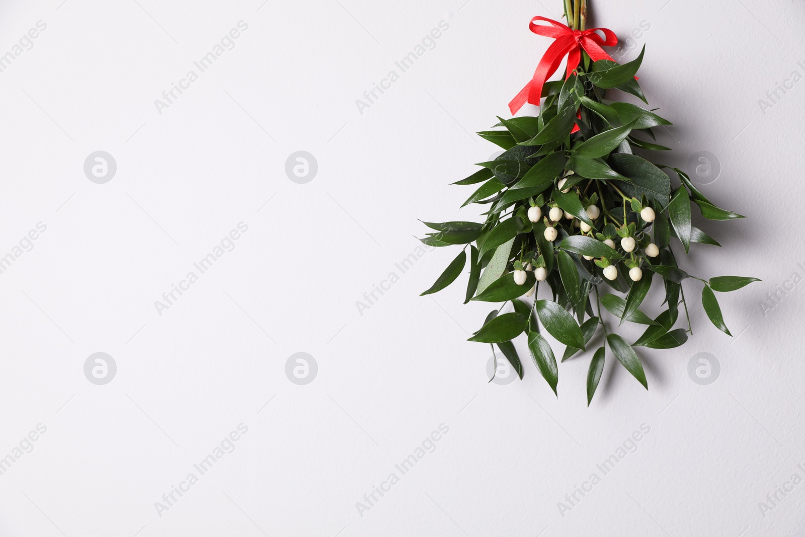 Photo of Mistletoe bunch with red bow hanging on white wall, space for text. Traditional Christmas decor