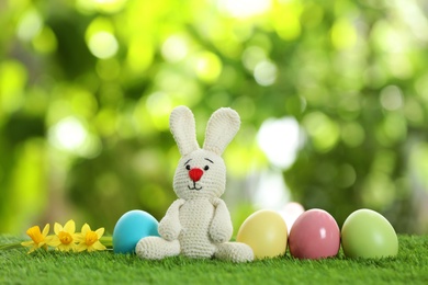 Photo of Cute Easter bunny toy and dyed eggs on green grass against blurred background, space for text