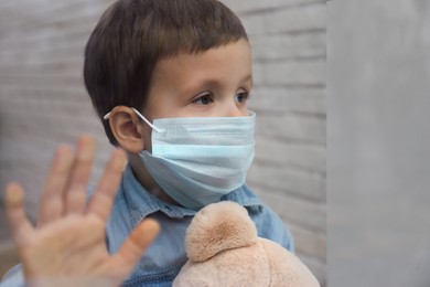 Photo of Sad little boy in protective mask looking out of window indoors, view from outside. Staying at home during coronavirus pandemic