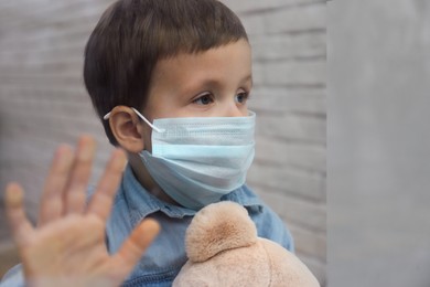 Sad little boy in protective mask looking out of window indoors, view from outside. Staying at home during coronavirus pandemic