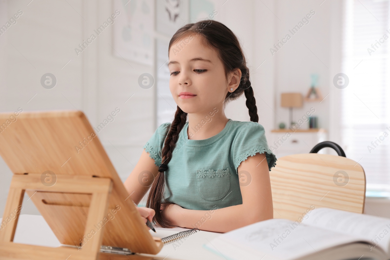 Photo of Little girl doing homework with tablet at table in room